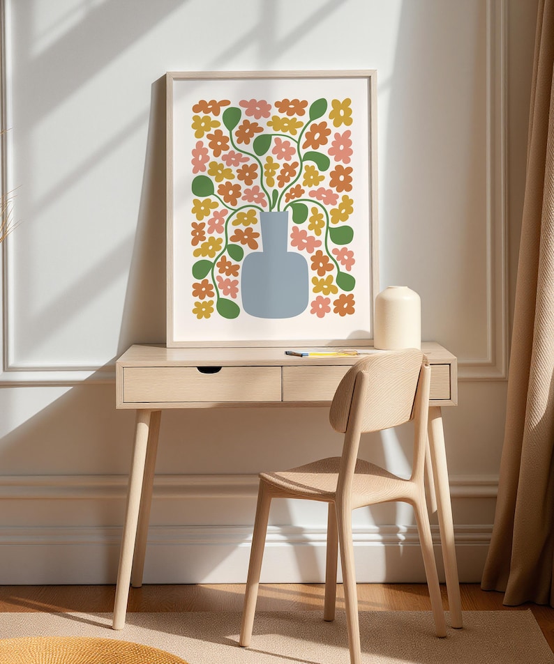 May include: A framed floral art print featuring a blue vase filled with colorful flowers in shades of orange, pink, and yellow. The artwork is displayed on a light wood desk with a matching chair, creating a modern and stylish home decor scene.