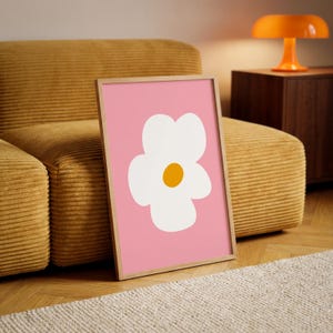 May include: A framed art print featuring a white flower with a yellow center on a pink background. The artwork is in a wooden frame and leans against a yellow corduroy sofa. A retro orange lamp sits on a dark wooden cabinet.