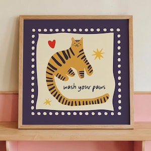 K&ouml;nnte beinhalten: Ein quadratischer gerahmter Wandkunstdruck mit einer gelben Katze mit schwarzen Streifen auf cremefarbenem Hintergrund. Die Katze ist von einem roten Herz, zwei gelben Sternen und dem Text "wash your paws" in Schwarz umgeben. Der Rand ist dunkelviolett mit wei&szlig;en Punkten.