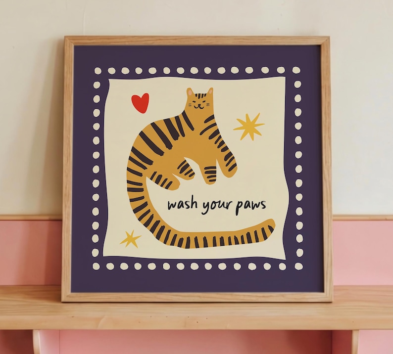 K&ouml;nnte beinhalten: Ein quadratischer gerahmter Wandkunstdruck mit einer gelben Katze mit schwarzen Streifen auf cremefarbenem Hintergrund. Die Katze ist von einem roten Herz, zwei gelben Sternen und dem Text "wash your paws" in Schwarz umgeben. Der Rand ist dunkelviolett mit wei&szlig;en Punkten.