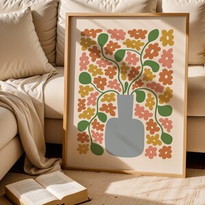 May include: A framed art print featuring a floral design. The artwork showcases a gray vase filled with stylized flowers in shades of orange, pink, and yellow, with green leaves. The print is set against a cream background and framed in light wood.