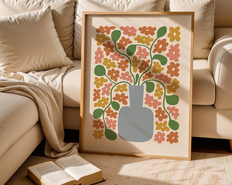 May include: A framed art print featuring a floral design. The artwork showcases a gray vase filled with stylized flowers in shades of orange, pink, and yellow, with green leaves. The print is set against a cream background and framed in light wood.
