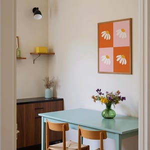 May include: A dining room scene featuring a light blue table and two wooden chairs with woven seats. A framed artwork with daisy designs hangs on the wall. A vase of flowers sits on the table, and a cabinet is visible.