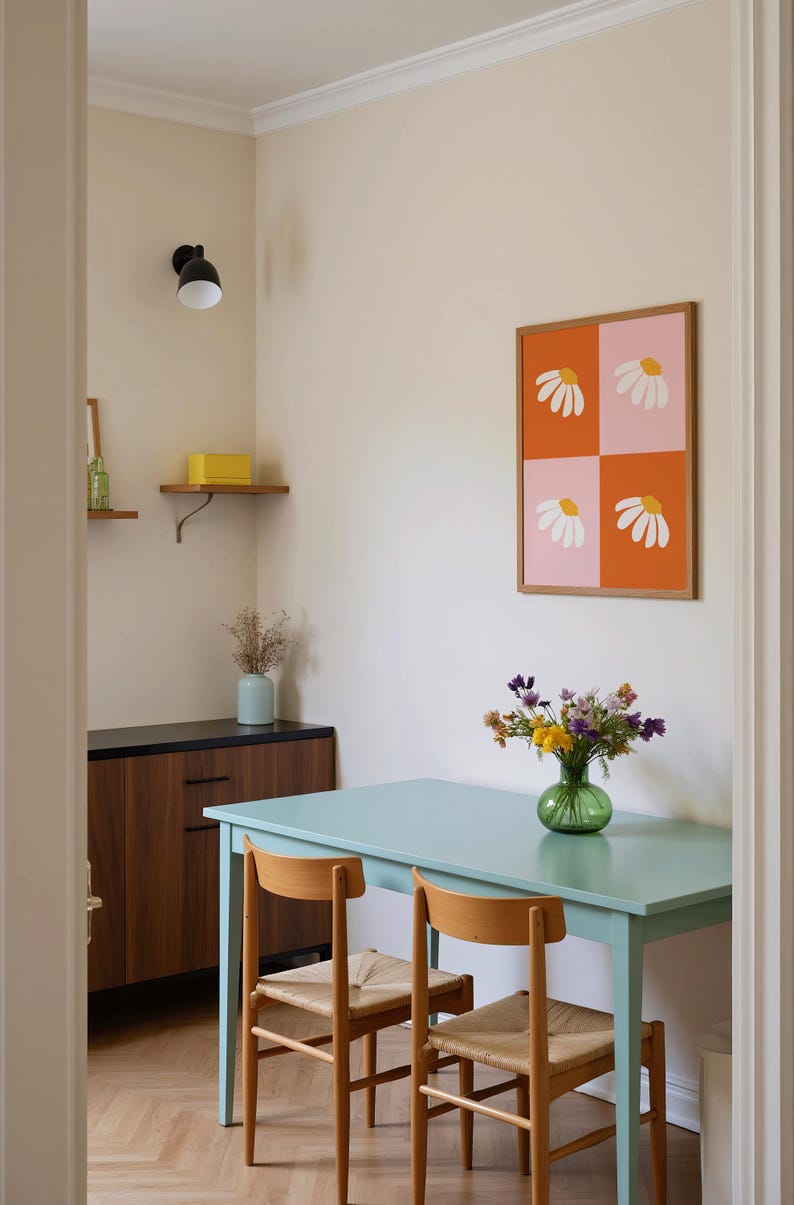 May include: A dining room scene featuring a light blue table and two wooden chairs with woven seats. A framed artwork with daisy designs hangs on the wall. A vase of flowers sits on the table, and a cabinet is visible.