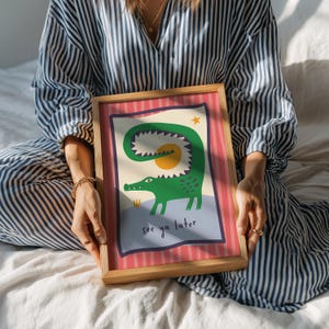 Pu&ograve; includere: Una stampa rettangolare incorniciata raffigurante un'illustrazione di un coccodrillo verde con la coda curva, un sole giallo e una piccola stella. Il testo "see ya later" &egrave; stampato in nero in basso. La stampa ha un bordo a righe rosa e rosse.