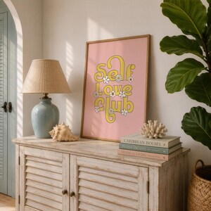 May include: A framed rectangular wall art print with a pink background featuring the yellow text "SELF LOVE CLUB" in a stylised font, accented with small white daisy illustrations, displayed on a wooden cabinet next to a lamp and books.