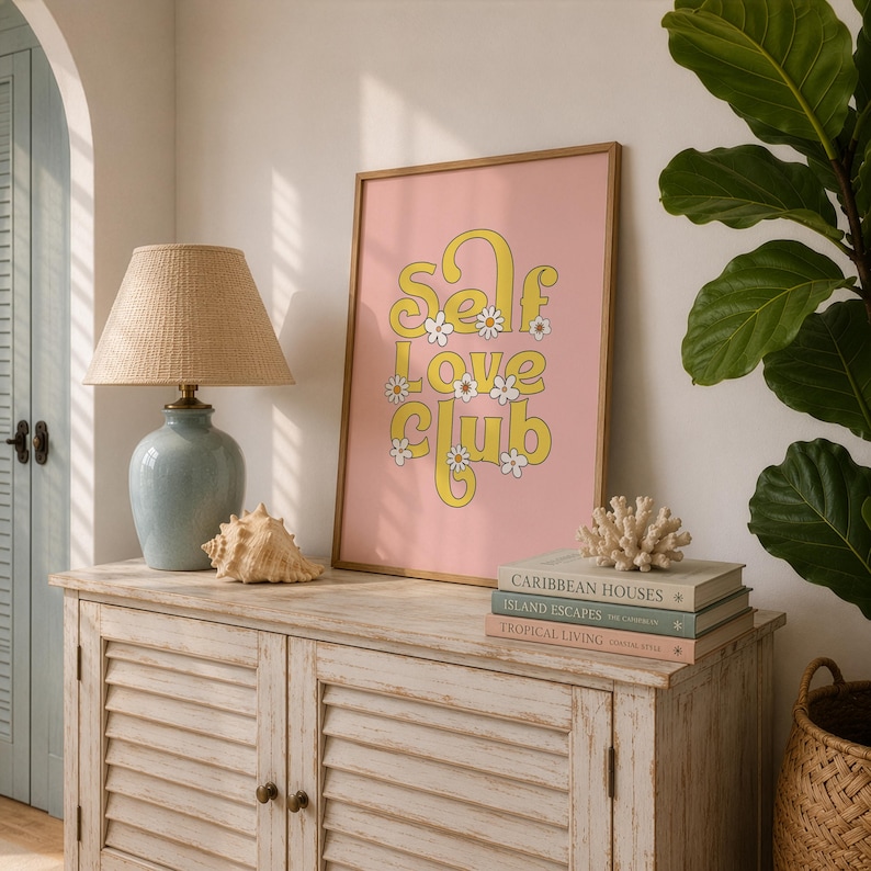 May include: A framed rectangular wall art print with a pink background featuring the yellow text "SELF LOVE CLUB" in a stylised font, accented with small white daisy illustrations, displayed on a wooden cabinet next to a lamp and books.