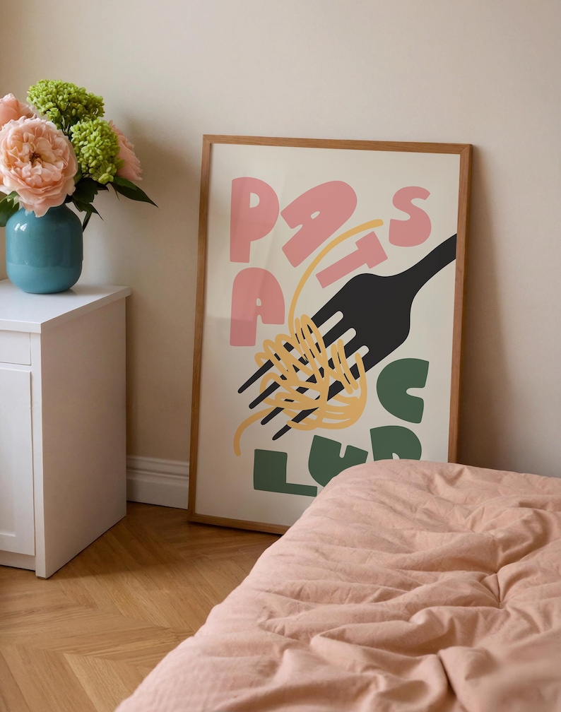 May include: A framed art print with the word "PASTA" in pink and green letters, a black fork, and yellow spaghetti. The artwork is displayed on a wall, with a bed and flowers in the foreground.