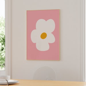 May include: A framed art print featuring a large white flower with a yellow center on a pink background. The artwork is displayed on a white wall in a room with natural light. The frame is gold-colored.