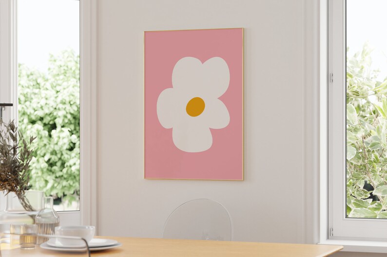 May include: A framed art print featuring a large white flower with a yellow center on a pink background. The artwork is displayed on a white wall in a room with natural light. The frame is gold-colored.
