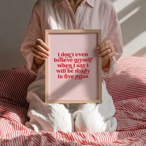 K&ouml;nnte beinhalten: Ein rechteckiger Kunstdruck in einem hellen Holzrahmen mit einem blassrosa Hintergrund und kr&auml;ftigem rotem Text, der lautet: I don't even believe myself when I say I will be ready in five mins. Die Typografie ist mit dekorativen Schn&ouml;rkeln versehen.