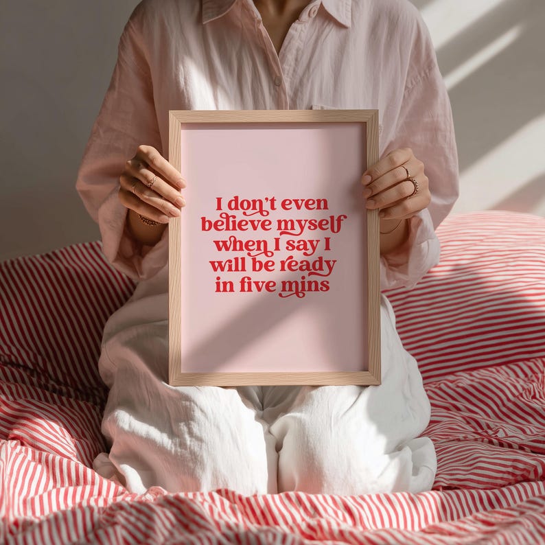 K&ouml;nnte beinhalten: Ein rechteckiger Kunstdruck in einem hellen Holzrahmen mit einem blassrosa Hintergrund und kr&auml;ftigem rotem Text, der lautet: I don't even believe myself when I say I will be ready in five mins. Die Typografie ist mit dekorativen Schn&ouml;rkeln versehen.