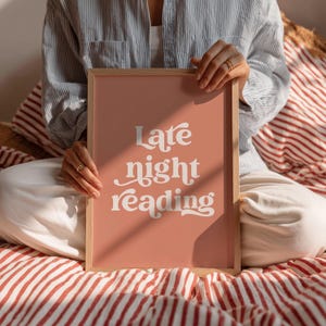 May include: A framed art print with the words "Late night reading" in white script on a peach background. The frame is light brown. The print is held by a person sitting on a bed with red and white striped bedding.