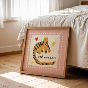 K&ouml;nnte beinhalten: Ein quadratisches gerahmtes Wandbild mit einem hellrosa Rand und wei&szlig;en Punkten. In der Mitte ist ein gelber Tiger mit schwarzen Streifen, ein kleines rotes Herz, zwei gelbe Sterne und der schwarze Text "wash your paws" zu sehen.
