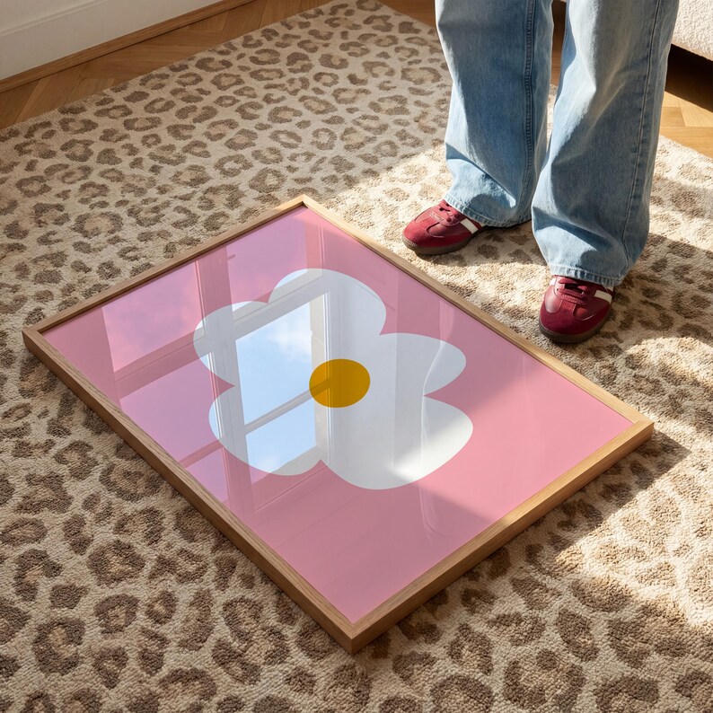 May include: A framed art print featuring a large white flower with a yellow center on a pink background. The artwork is displayed in a light brown wooden frame. The print is resting on a leopard print rug.