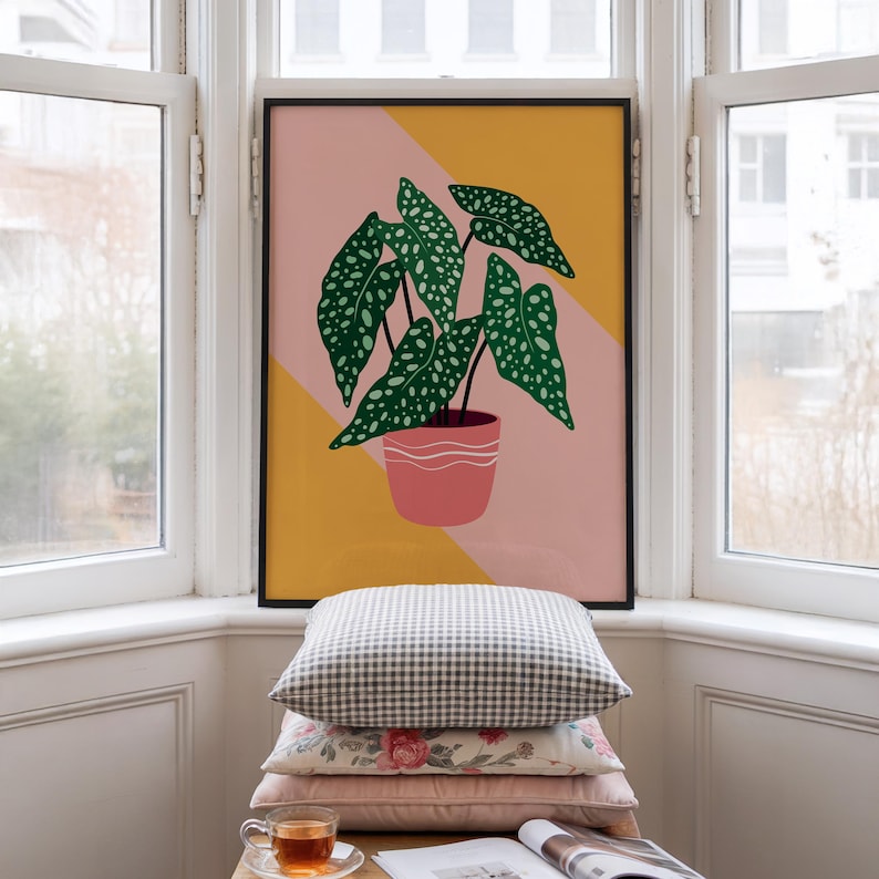 May include: A framed art print depicting a houseplant with green, spotted leaves in a pink pot. The background is a geometric design in pink and yellow. The artwork is displayed in a bright room, with a stack of cushions in front of it.