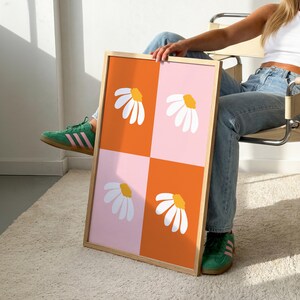 May include: A framed art print featuring a daisy design. The artwork is divided into four squares, alternating between orange and pink backgrounds. Each square displays a white daisy with a yellow center. The frame is made of light-colored wood.