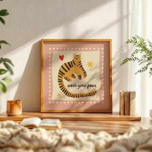 K&ouml;nnte beinhalten: Ein quadratischer gerahmter Kunstdruck mit einem stilisierten orange-schwarz gestreiften Tiger auf cremefarbenem Hintergrund mit rosa Rand. Die Illustration enth&auml;lt ein kleines rotes Herz, zwei gelbe Sterne und den Text wash your paws in Schwarz.