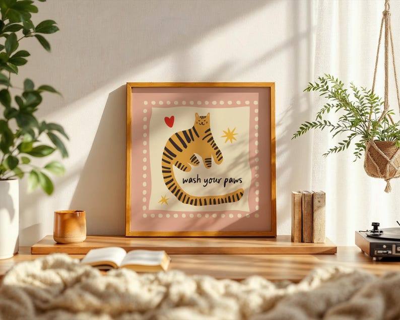 K&ouml;nnte beinhalten: Ein quadratischer gerahmter Kunstdruck mit einem stilisierten orange-schwarz gestreiften Tiger auf cremefarbenem Hintergrund mit rosa Rand. Die Illustration enth&auml;lt ein kleines rotes Herz, zwei gelbe Sterne und den Text wash your paws in Schwarz.