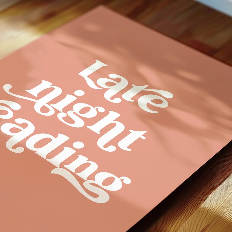 May include: A pink poster with the words "Late night reading" in a white, retro font. The poster is angled, with a wooden surface visible in the foreground and background. The lighting suggests a warm, indoor setting.