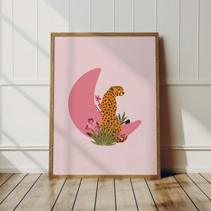 May include: A framed art print featuring a cheetah with orange and black spots, perched on a pink crescent moon. The cheetah is surrounded by pink flowers and green foliage against a light pink background. The artwork is in a wooden frame.