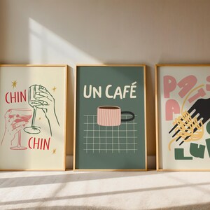 May include: Three framed art prints with gold frames. The prints feature illustrations of wine glasses, a coffee cup with the text "UN CAF&Eacute;", and pasta with a fork. The prints are in a modern, minimalist style.