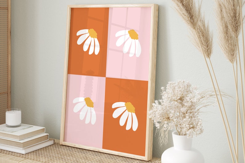 May include: A framed art print featuring a daisy design. The artwork is divided into four squares, alternating between orange and pink. Each square has a white daisy with a yellow center. The frame is light wood.