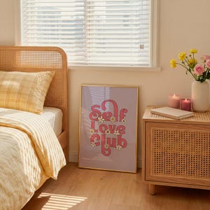 May include: A framed vertical wall art print featuring the text Self Love Club in a bold pink retro font, decorated with small white and yellow daisy illustrations, set against a light purple background in a gold-coloured metal frame.