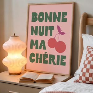 May include: A framed pink art print with the words "BONNE NUIT MA CH&Eacute;RIE" in green, along with a cherry illustration. A decorative lamp and an open book sit on a nightstand next to the bed.
