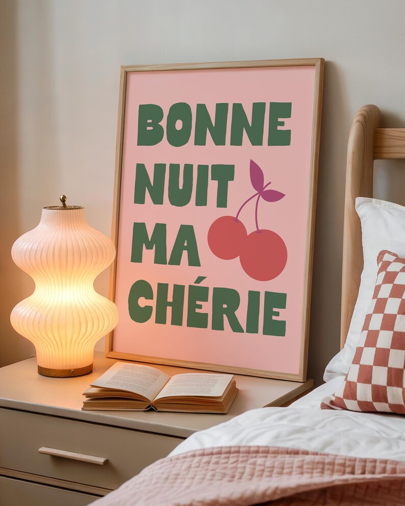 May include: A framed pink art print with the words "BONNE NUIT MA CH&Eacute;RIE" in green, along with a cherry illustration. A decorative lamp and an open book sit on a nightstand next to the bed.