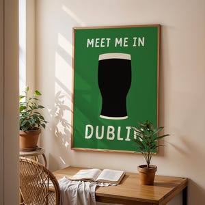 May include: A framed poster with a green background and the words "MEET ME IN DUBLIN" in white. A black pint of stout with a white head is centered on the poster. The poster is displayed on a wall above a wooden desk.