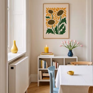 May include: A dining room scene featuring a framed sunflower art print, a white bookshelf with books, a blue vase with pink tulips, and a yellow candle. A white tablecloth covers the table, with a yellow teacup.