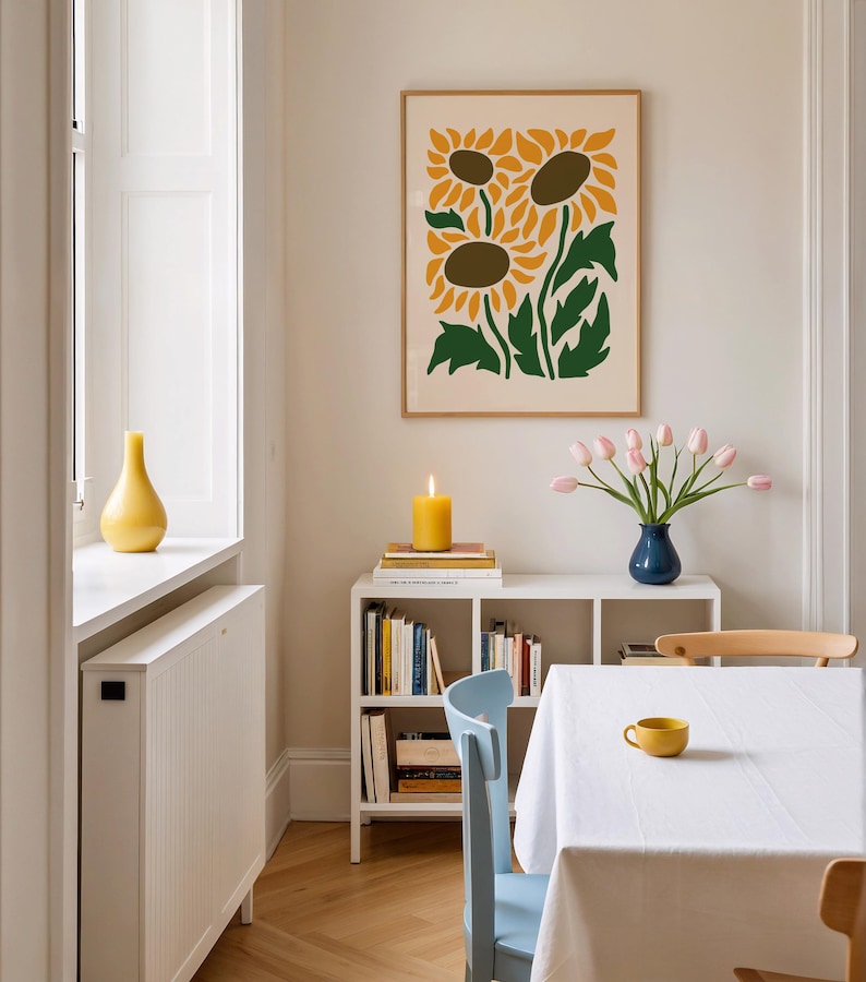 May include: A dining room scene featuring a framed sunflower art print, a white bookshelf with books, a blue vase with pink tulips, and a yellow candle. A white tablecloth covers the table, with a yellow teacup.