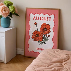 May include: A framed art print featuring a pink background with a white, wavy-edged center. The print displays the word "AUGUST" in red, along with illustrations of orange poppies and the word "poppy".