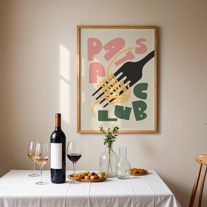 May include: A dining scene with a framed poster that reads "Pasta Club" in pink, black, and green. A bottle of red wine, wine glasses, and a wooden plate of food sit on a white tablecloth. A wooden chair is on either side of the table.