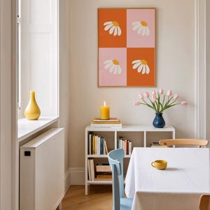 May include: A dining room scene featuring a framed artwork with daisy designs, a white bookshelf with books, a blue vase with pink tulips, and a yellow candle. A white tablecloth covers the table, with a yellow cup.