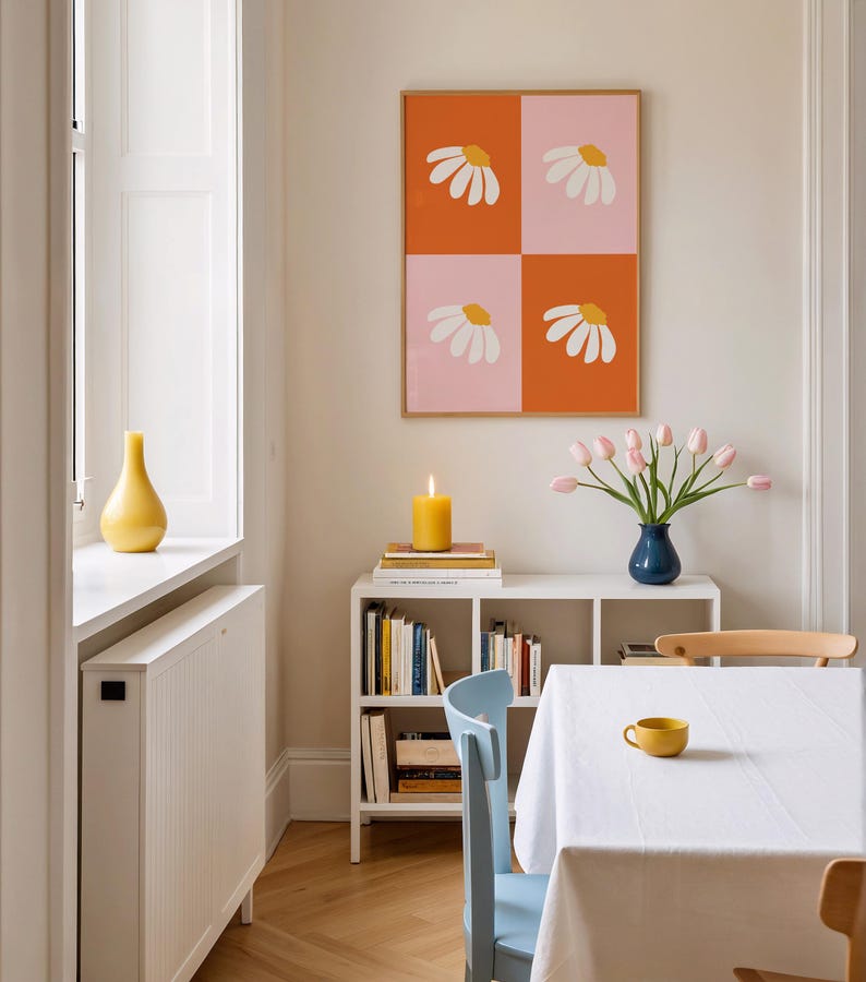 May include: A dining room scene featuring a framed artwork with daisy designs, a white bookshelf with books, a blue vase with pink tulips, and a yellow candle. A white tablecloth covers the table, with a yellow cup.