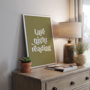 May include: A framed print with the words "Late night reading" in white script on an olive green background. The print is displayed on a wooden dresser, alongside a small potted succulent, a lamp, and a vase of flowers.