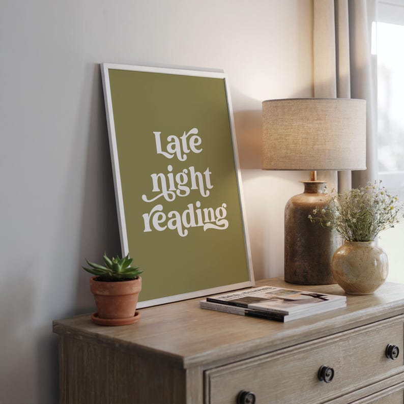 May include: A framed print with the words "Late night reading" in white script on an olive green background. The print is displayed on a wooden dresser, alongside a small potted succulent, a lamp, and a vase of flowers.