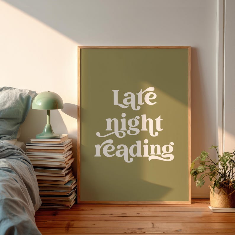 May include: A framed print with the words "Late night reading" in white script on an olive green background. The print is displayed on a wooden floor, next to a bed with a green lamp and a stack of books.
