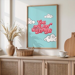 May include: A light blue poster with a white frame. The poster has a pink and white text that says "You are my Sunshine" with white clouds and a palm tree shadow.