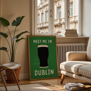 May include: A framed green poster with the text "MEET ME IN DUBLIN" and a graphic of a pint of stout. The poster is in a living room with a beige sofa, wooden chair, and a large potted plant. A window with a view of a building is in the background.
