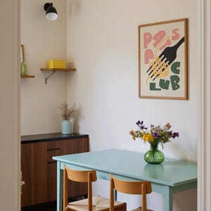 May include: A dining room scene with a light blue table and two wooden chairs. A framed poster with the words "Pasta Club" hangs on the wall. A vase of flowers sits on the table, and a wooden cabinet is visible.