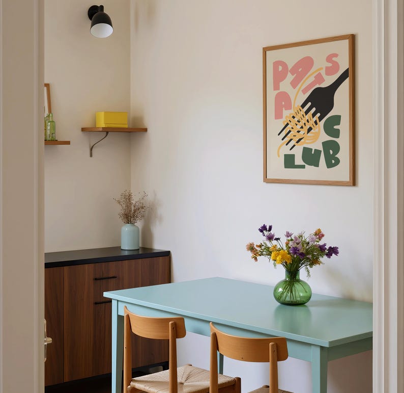 May include: A dining room scene with a light blue table and two wooden chairs. A framed poster with the words "Pasta Club" hangs on the wall. A vase of flowers sits on the table, and a wooden cabinet is visible.