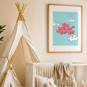 May include: A white teepee tent with a wooden frame, a white crib with a beige blanket, and a framed print with a blue background and the text "You are my sunshine" in pink and white.