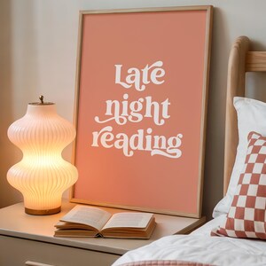 May include: A framed art print with the words "Late night reading" in white script against a coral background. A glowing, ribbed glass lamp and an open book sit on a nightstand. A bed with a patterned pillow is in the background.
