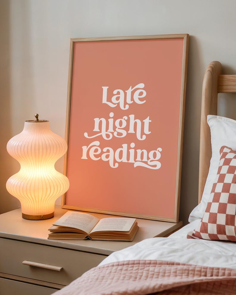 May include: A framed art print with the words "Late night reading" in white script against a coral background. A glowing, ribbed glass lamp and an open book sit on a nightstand. A bed with a patterned pillow is in the background.