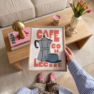 May include: A framed art print with the words "CAFE CON LECHE" in orange letters. The print features a gray coffee maker and a blue coffee cup. The image is held by a person wearing a blue and white striped shirt.