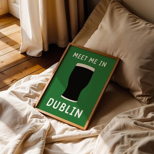 May include: A framed art print with a green background and the words "MEET ME IN DUBLIN" in white. The image features a black pint of stout. The frame is a light brown color. The print is resting on a bed with a beige comforter and pillow.