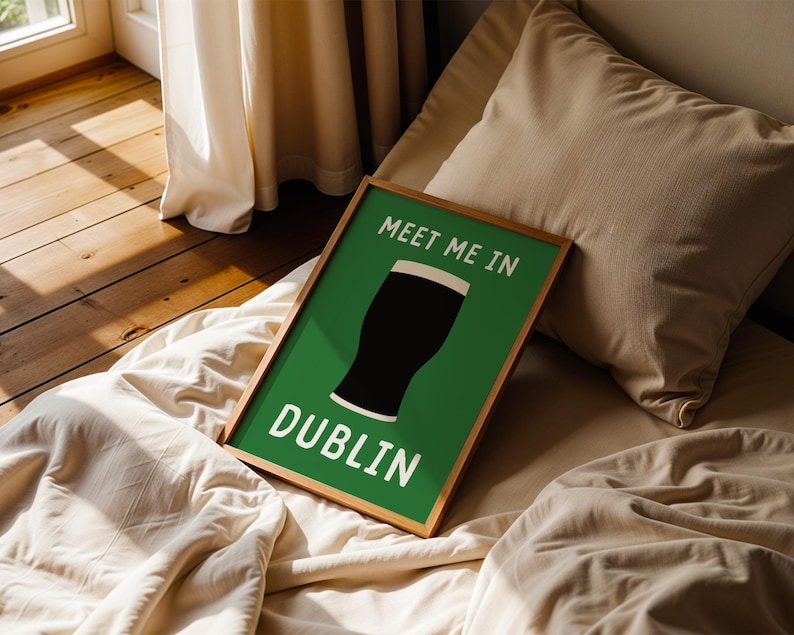 May include: A framed art print with a green background and the words "MEET ME IN DUBLIN" in white. The image features a black pint of stout. The frame is a light brown color. The print is resting on a bed with a beige comforter and pillow.